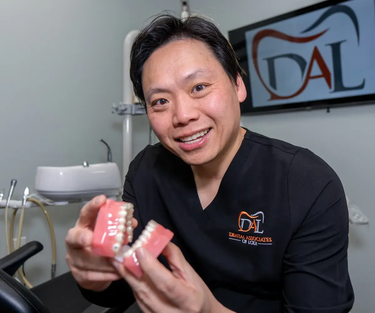 A dentist holding a dental model, smiling, inside a dental office with equipment visible in the background.