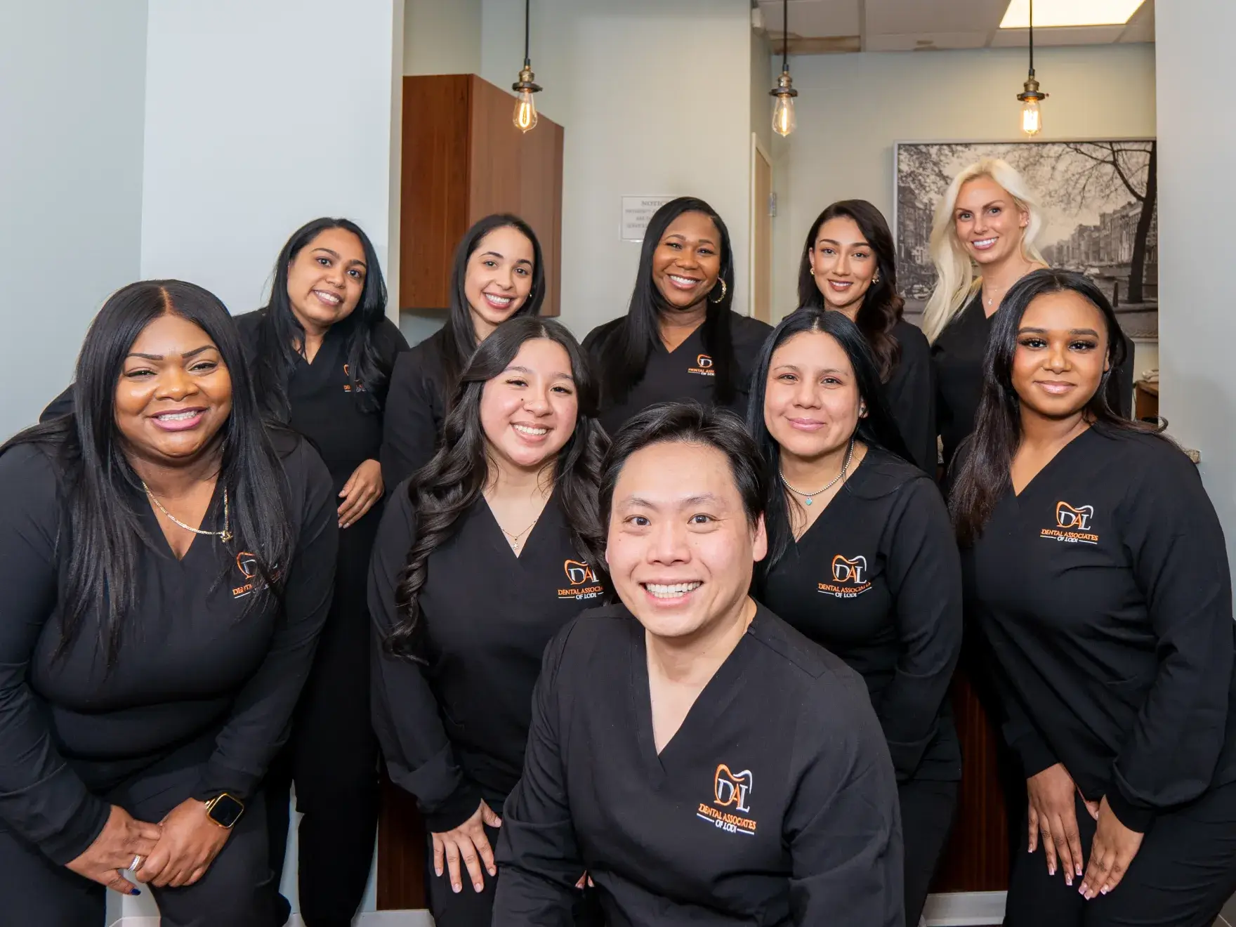 Group of ten diverse dental professionals wearing black scrubs with Dental Associates of Lodi logo, smiling indoors.