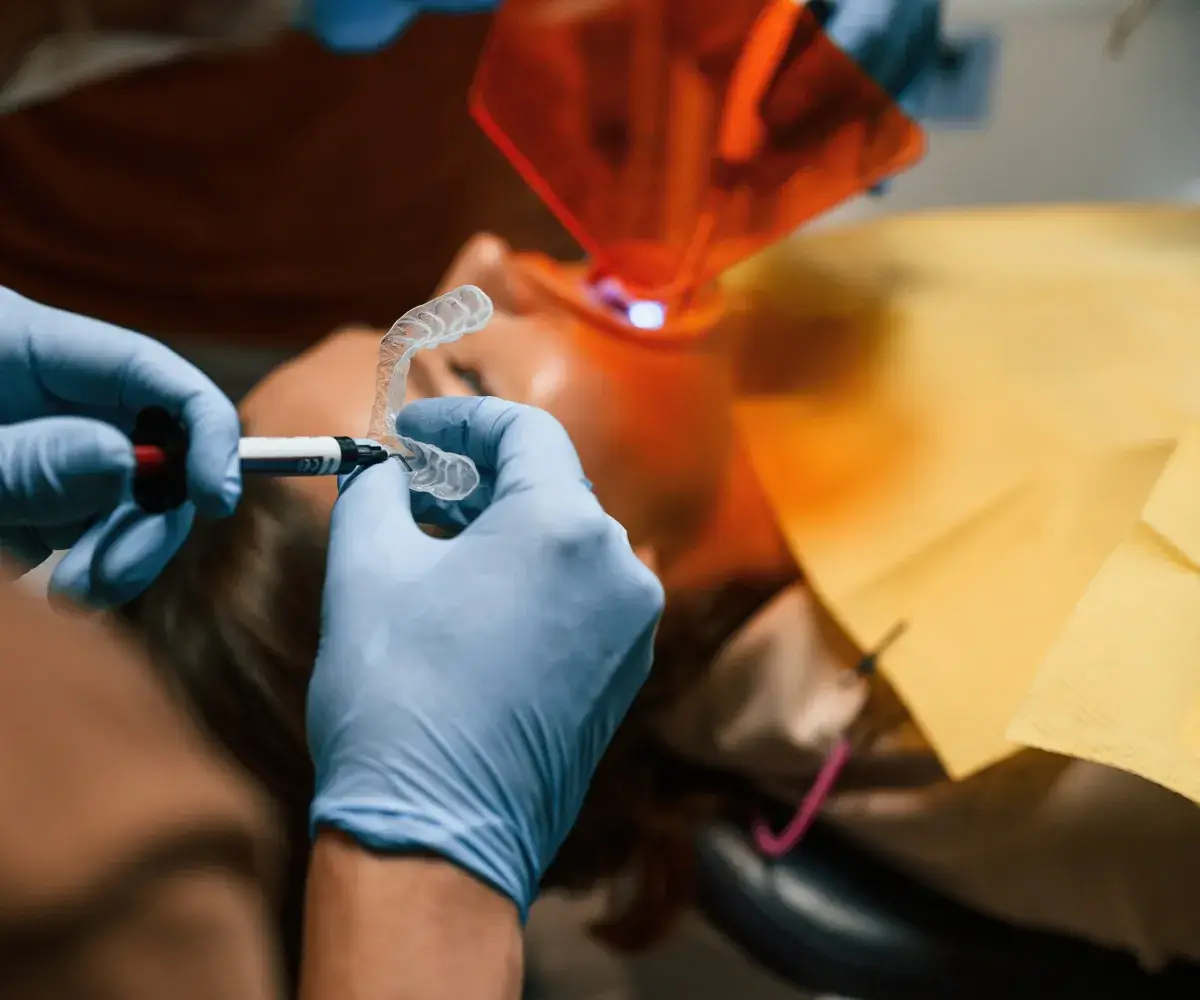 Dentist wearing gloves applies gel to a dental mold in front of a patient.