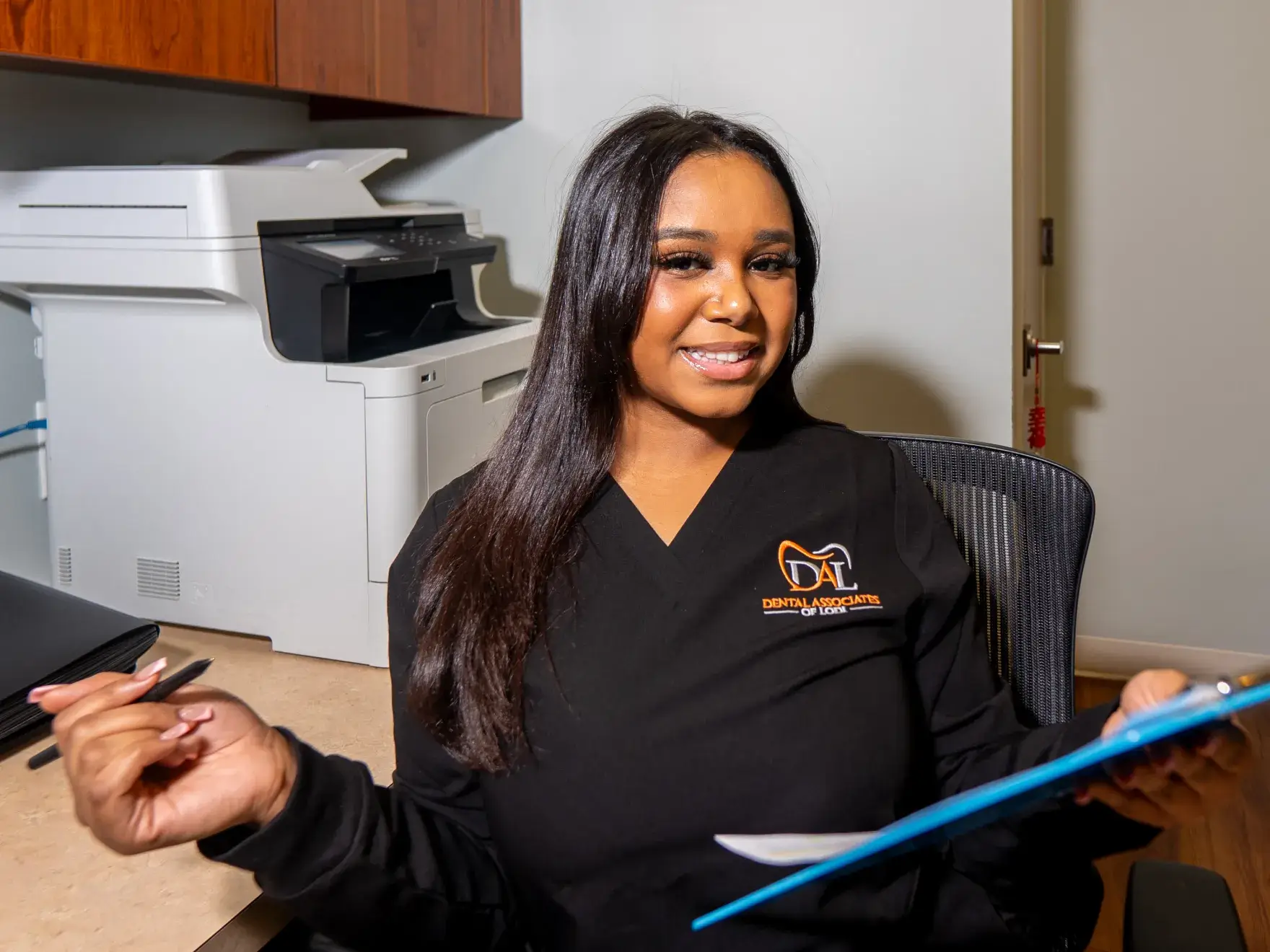 A woman in black scrubs sits at a desk holding a clipboard, smiling at the camera.