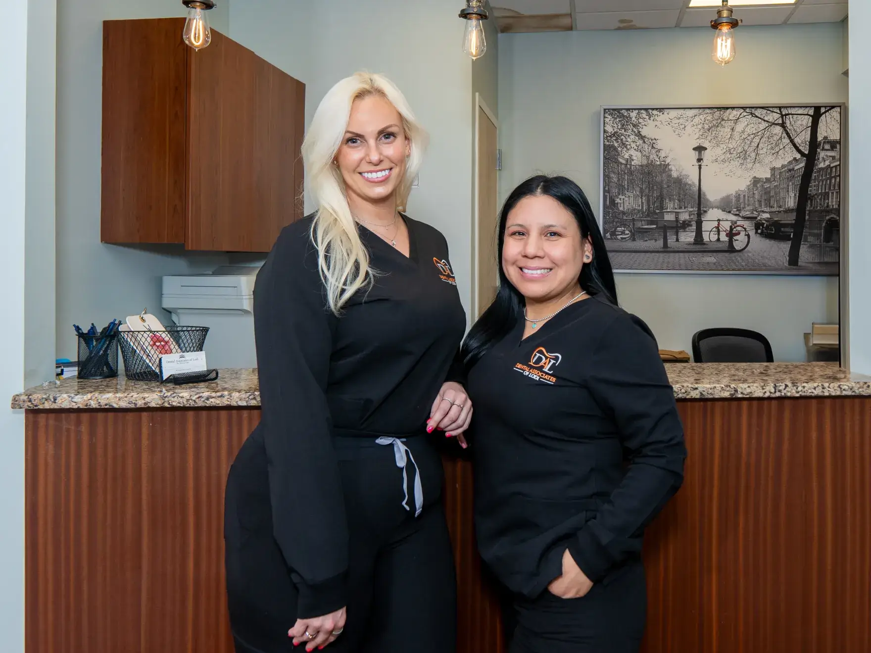 Two women in matching black uniforms stand smiling in front of a reception desk.